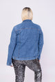 Modelo Victoria está de costas e vestindo jaqueta jeans basik na cor azul, peça possui bolsos na frente e botões de metal.