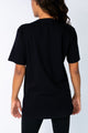 Modelo Nathália está de costas e usando camiseta bordada raio preta. Peça possui bordado manual de paetês somente na frente.