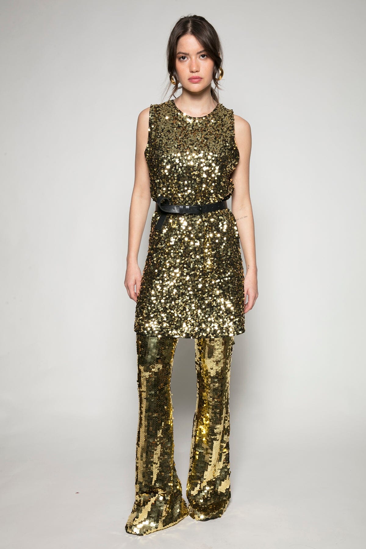 Modelo Ana usa calça flare sole dourada e vestido mini luna dourado com um cinto localizado na cintura. Peças bordadas à mão.