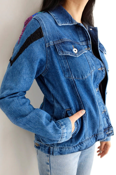 Modelo Katy está vestindo jaqueta jeans logus colorida. Peça com fechamento de botões de metal na frente, bolsos e mangas.