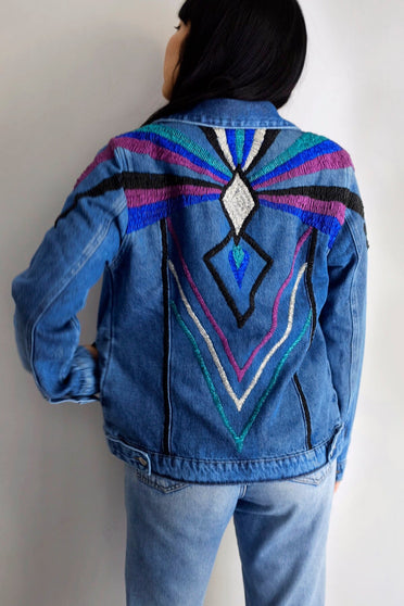 Modelo Katy está de costas e usando jaqueta jeans logus colorida. Peça com bordado manual de miçangas localizado nas costas.