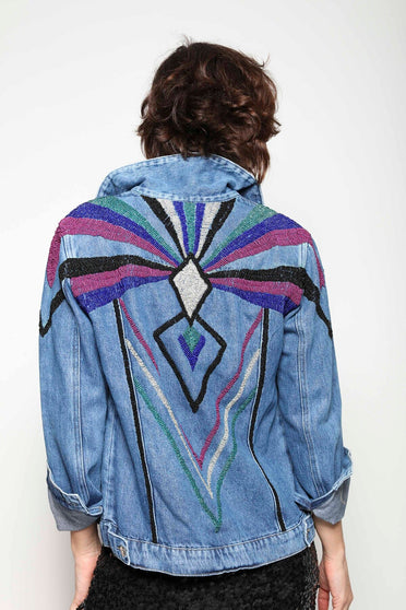 Modelo Isabel está de costas e vestindo jaqueta jeans bordada logus na cor colorido. Peça com bordado de miçangas nas costas.
