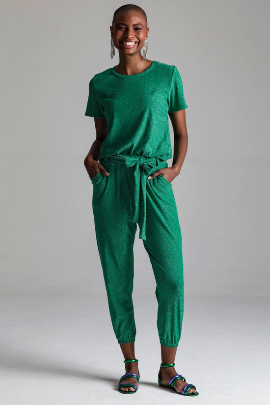 Modelo em pé está usando calça jogger luxor verde. Peça confortável com tecido leve de malha lurex verde. Possui muito brilho