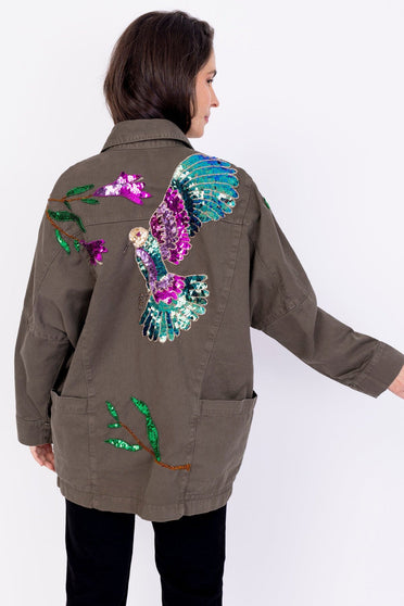Modelo Simone está de costas e usa jaqueta parka beija-flor. Peça mais larga com um estilo oversize, possui bordado manual.