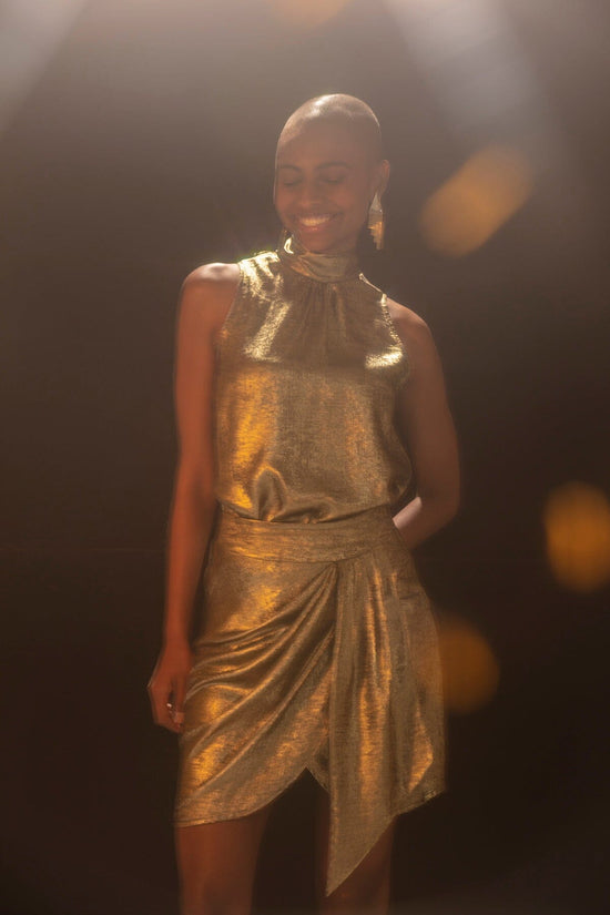 Modelo em pé sorri enquanto usa conjunto Atenas dourado. Regata de gola alta com brilho metalizado. Modelagem mais soltinha.
