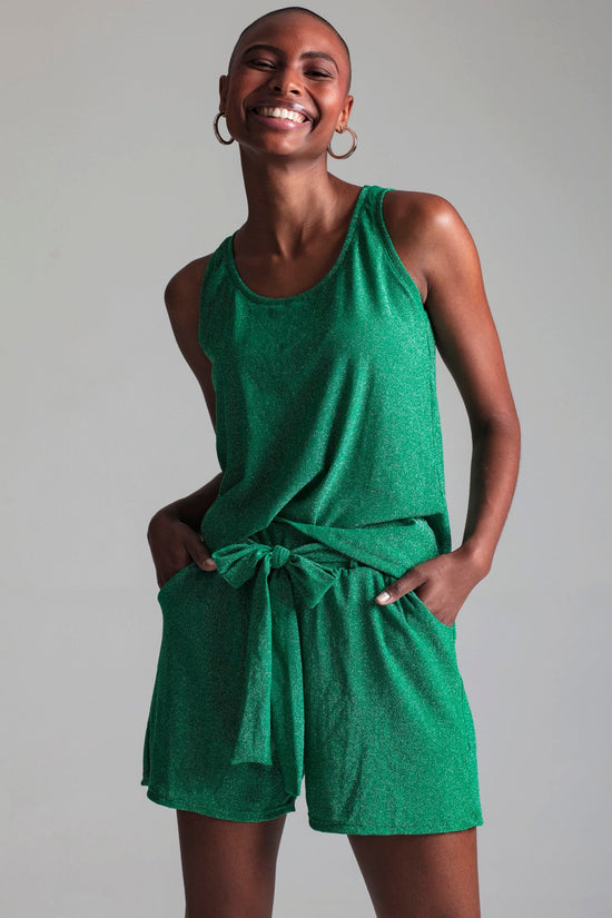 Modelo em pé usa shorts Luxor Joulik na cor verde. Peça confortável em malha lurex de base verde e muito brilho na cor prata.
