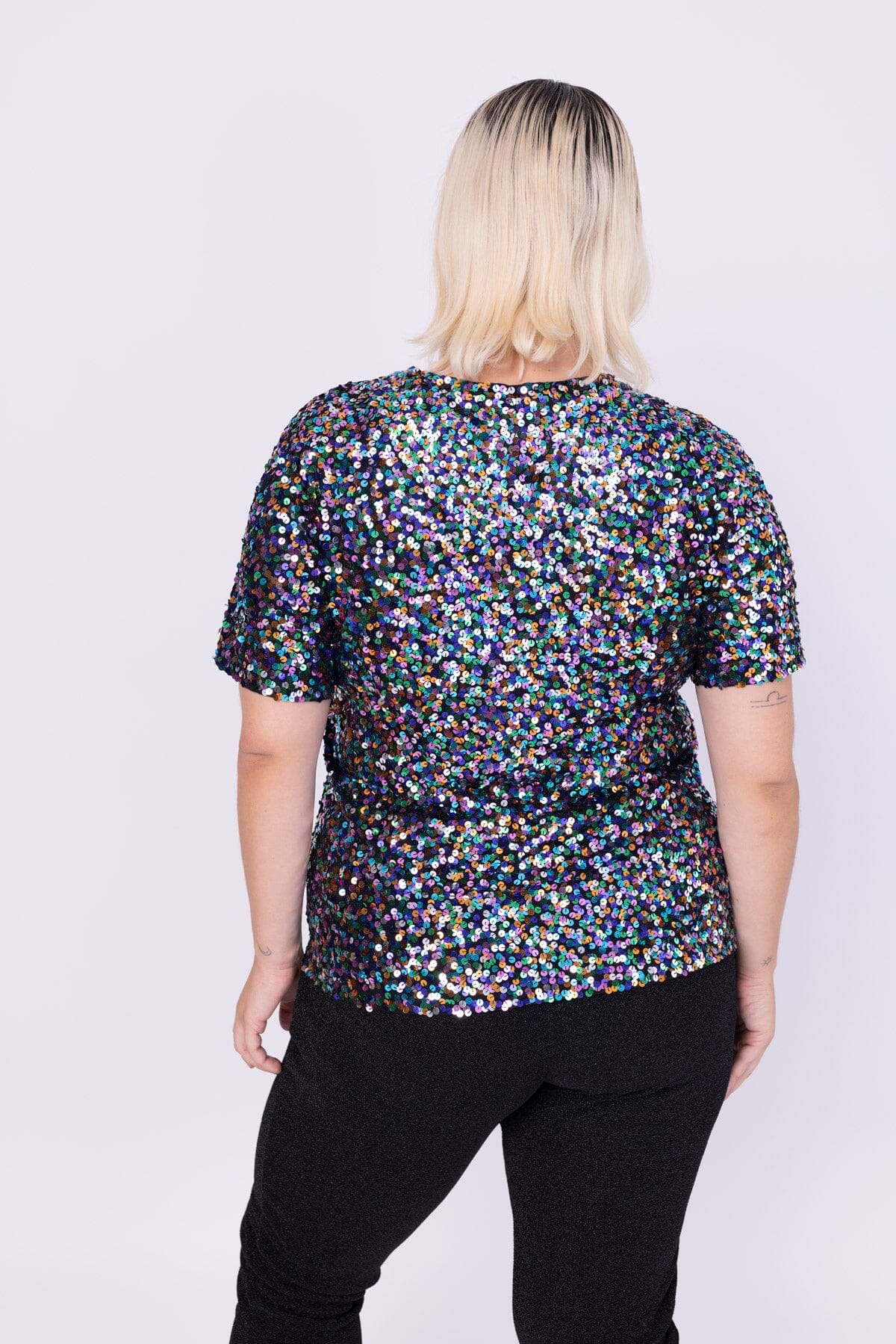 Modelo Victoria está de costas e usa blusa luna. Blusa com muito brilho de paetês coloridos bordados à mão. Feito no Brasil.