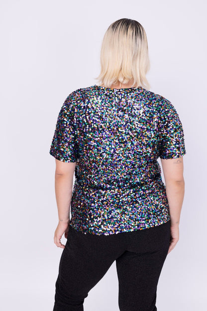 Modelo Victoria está de costas e usa blusa luna. Blusa com muito brilho de paetês coloridos bordados à mão. Feito no Brasil.