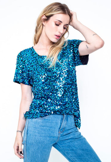 Modelo Juliana está com a mão na cabeça, ela veste a blusa luna de manga curta, peça bordada á mão com paetês na cor turquesa